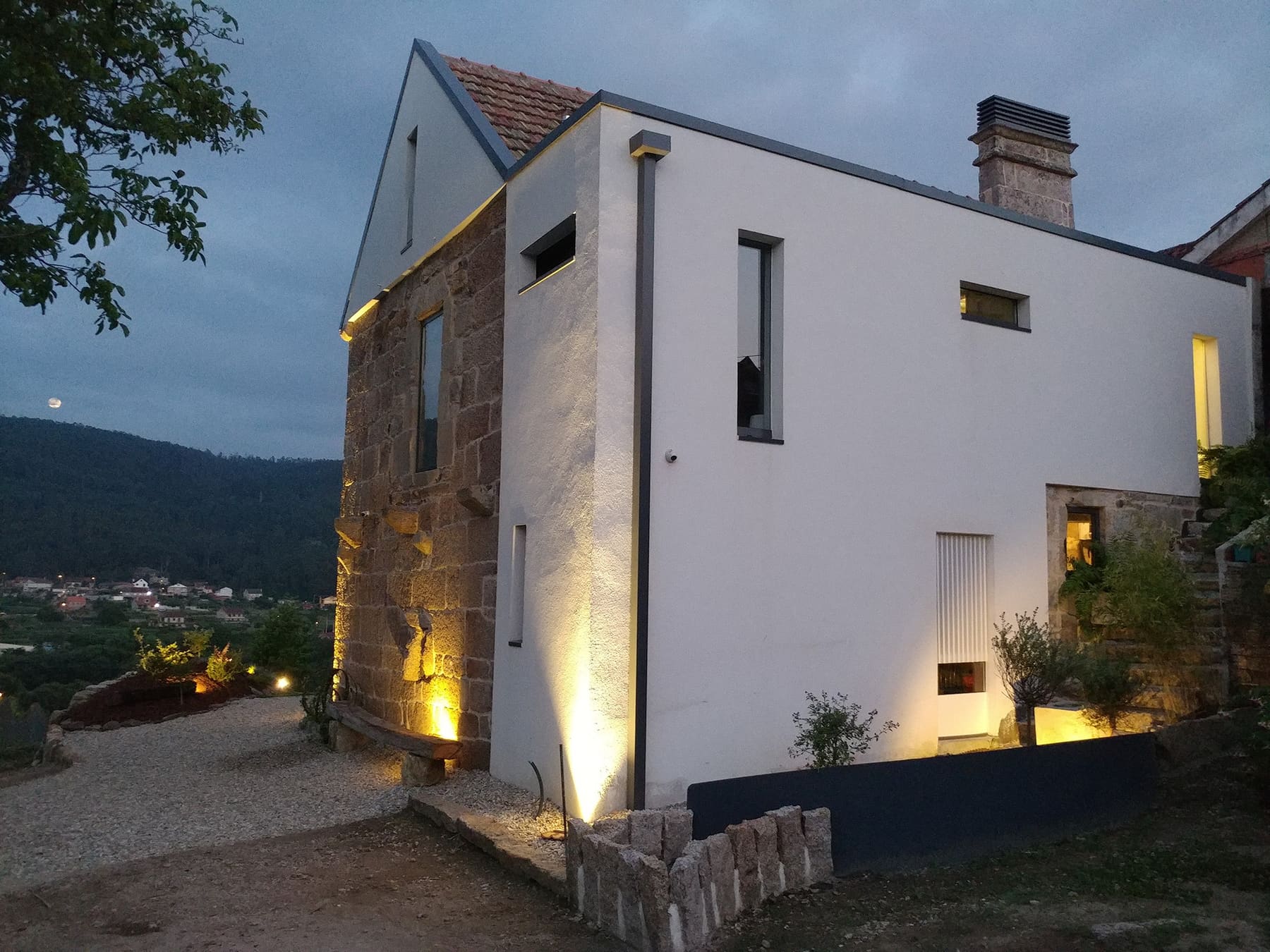 Casa dos Parladoiros al anochecer: el hastial de granito original junto al volumen blanco de la restauración, con luz rasante y el valle al fondo.
